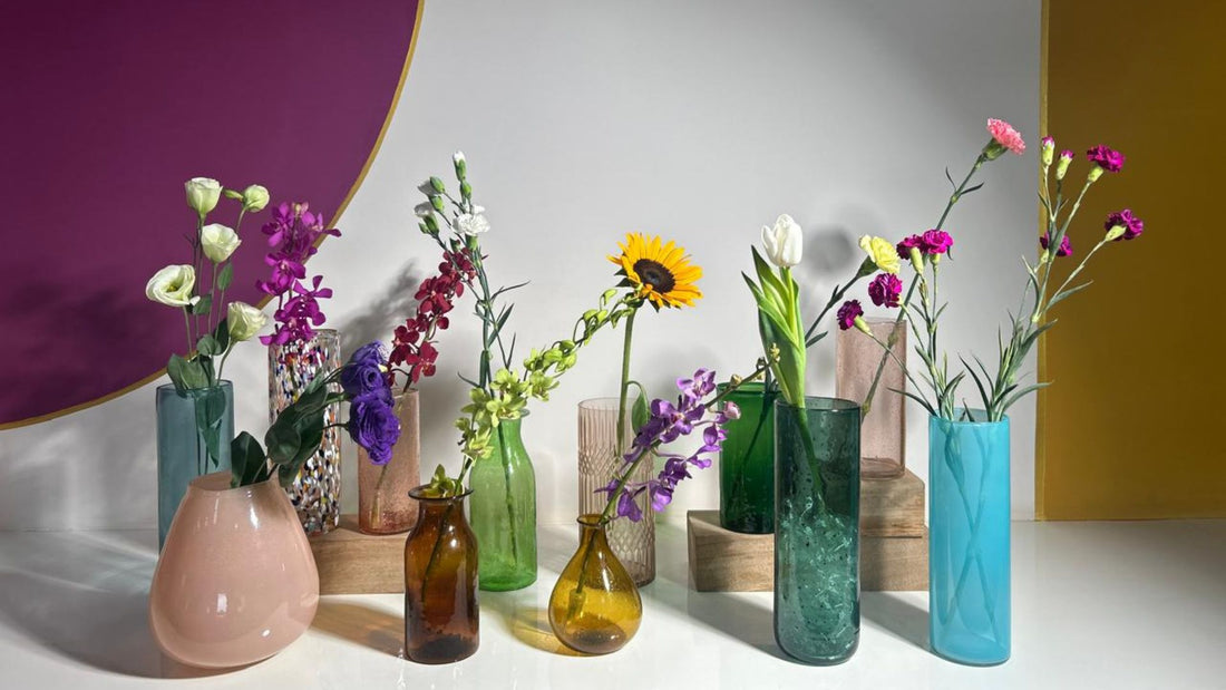 Colorful fresh flowers arranged in various uniquely shaped glass vases, including sunflowers, tulips, carnations, and orchids, displayed on a modern table with a vibrant purple and yellow backdrop.