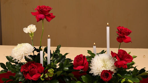 Elegant floral arrangement featuring red roses, white chrysanthemums, and white candles on a table with a beige tablecloth.