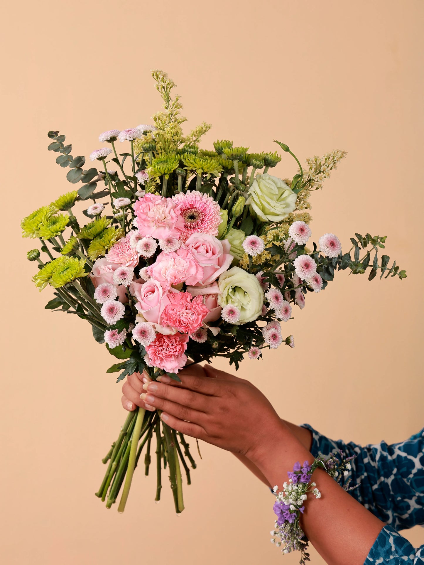 A hand presents a bouquet of Sweet Avalanche roses, pink carnations, pink gerberas, green chrysanthemums, green eustoma, and light calimero, complemented by a matching floral wristlet