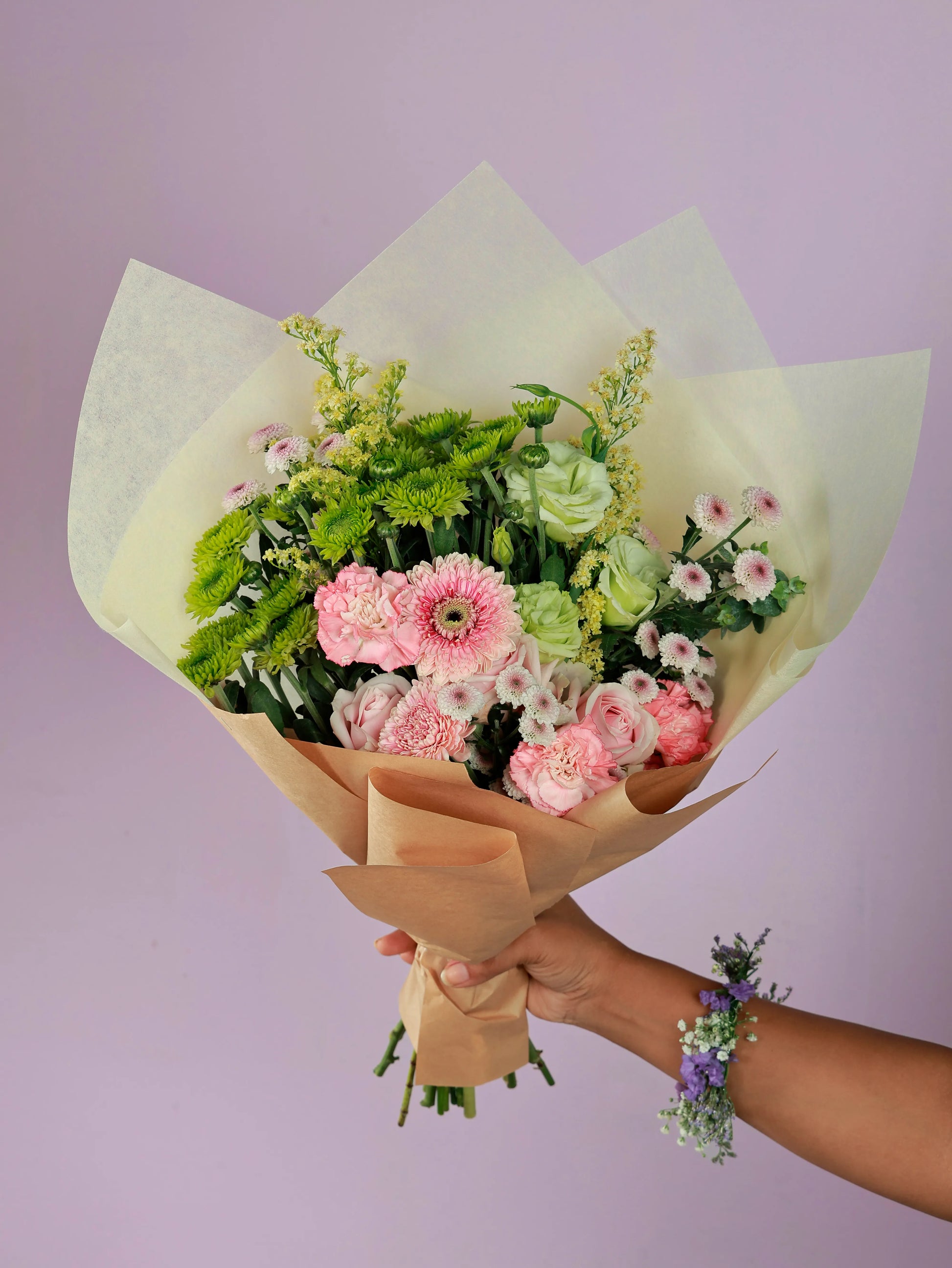 Ready-to-gift bouquet of Sweet Avalanche roses, pink carnations, pink gerberas, green chrysanthemums, green eustoma, and light calimero, professionally wrapped and held by a hand with a coordinating wrist corsage