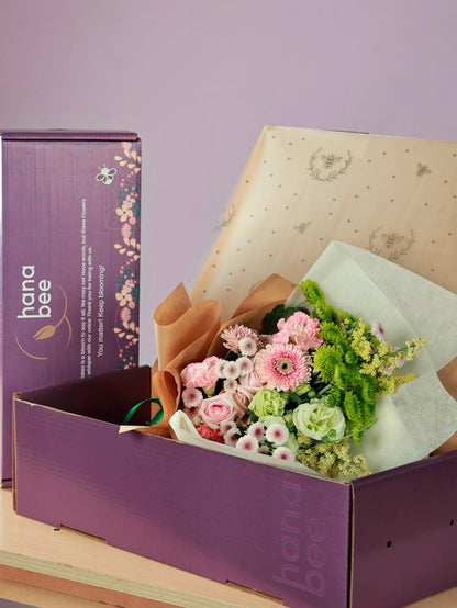 A wrapped bouquet of Sweet Avalanche roses, pink carnations, pink gerberas, green chrysanthemums, green eustoma, and light calimero packaged in a Hanabee box, ready for shipping or delivery.