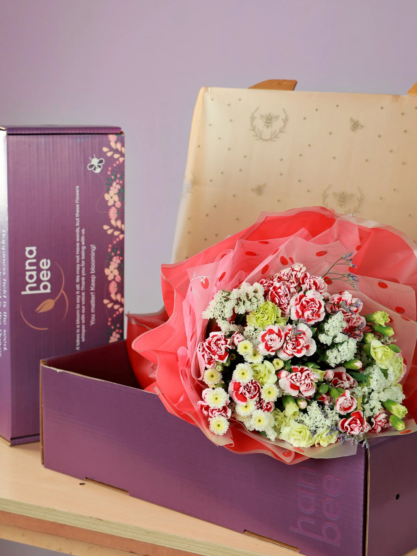 Red, white, and cream flower bouquet wrapped in cellophane, presented in a purple Hana Bee branded gift box